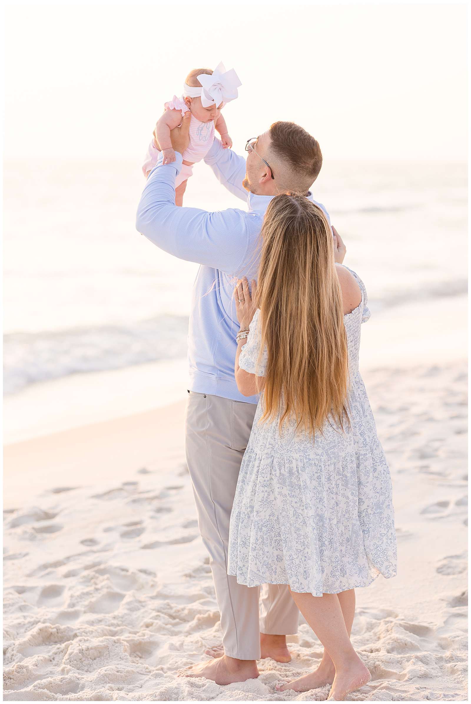 Couple stands on the beach with their back towards the camera as new Dad holds his baby girl up in the air as her parents look up at her.  Click to see more of this Rosemary Beach session with Rebecca Rice.  - rebeccaricephoto.com