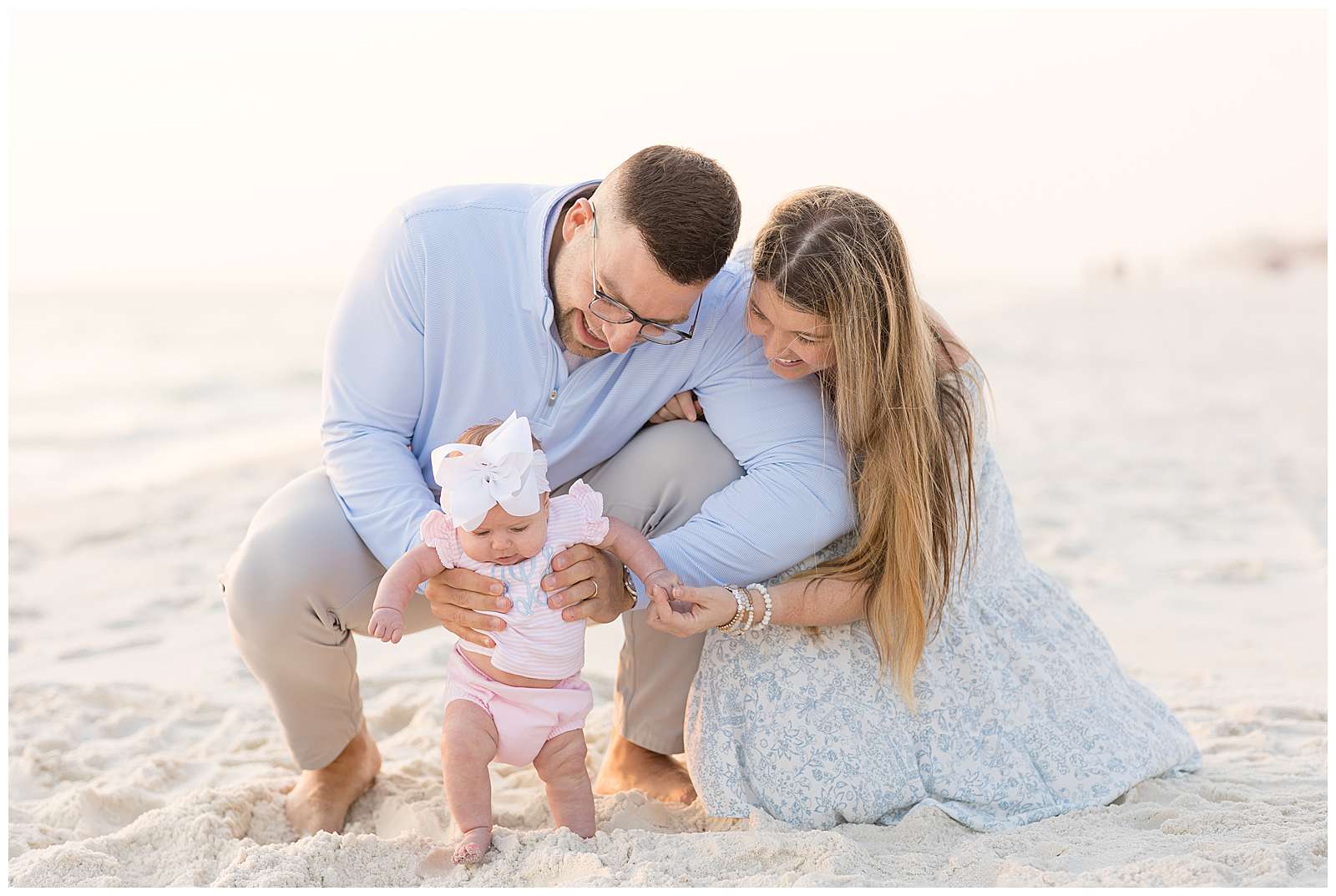 New parents of baby girl squat down on Rosemary Beach together and new Dad, holds his baby girl up as she puts her toes in the sand.  The baby wears a pink and white striped outfit with her initials monogrammed on the front and a big, white, headband bow.  See more of this adorable, Rosemary Beach family portrait session on the blog! -rebeccaricephoto.com