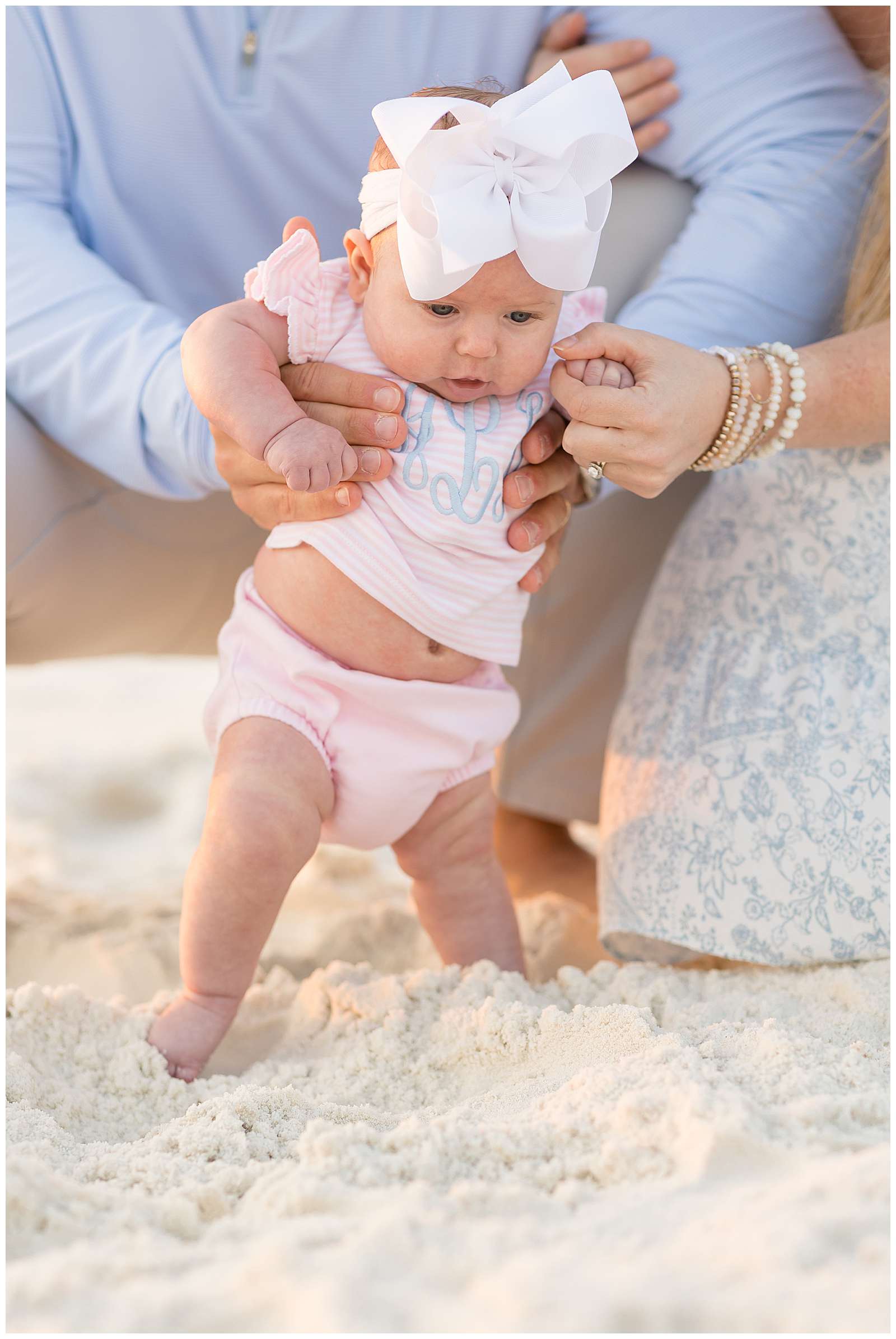 Sweet baby girl who wears a monogrammed, pink and white stripped outfit, puts her toes in the sand and is being held up by her Dad and Mom who we see their hands in the image.  Click to see more of this Rosemary Beach family session from Rebecca Rice Photo! - rebeccaricephoto.com