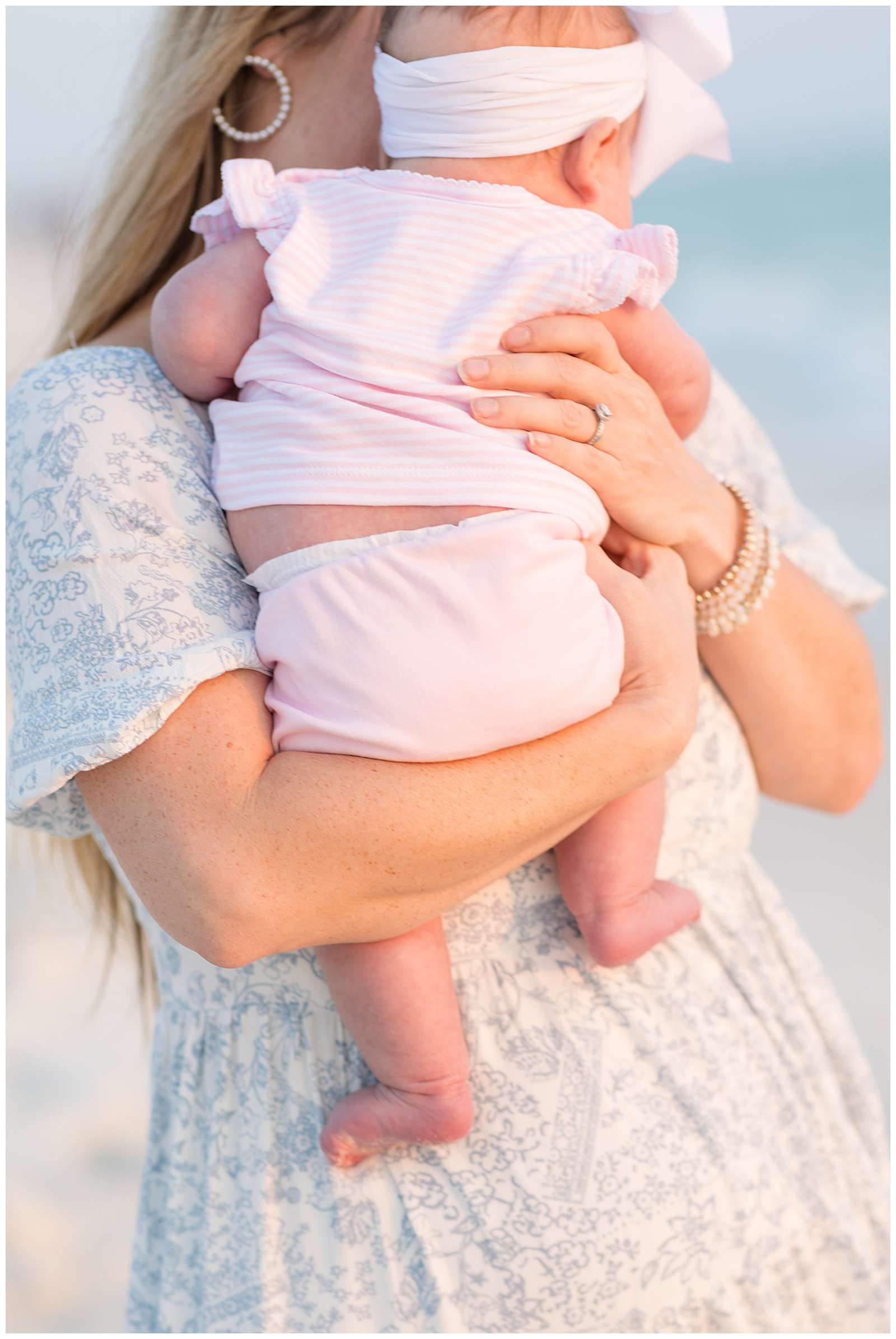 Close-up, detailed image shows the back of baby girl who is wearing a pink and white stripped outfit being held by her mother's arm and hand during their Rosemary Beach family portrait session. - rebeccaricephoto.com