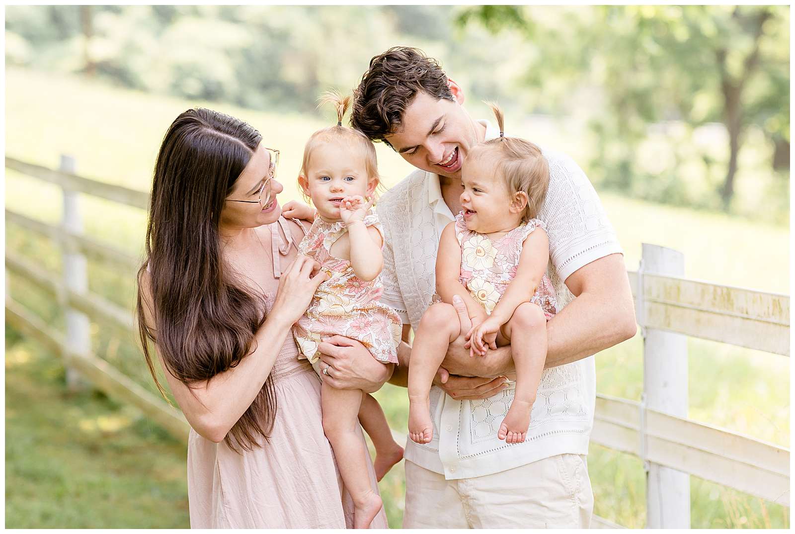 Family with 1 year old twins stand in front of a white fence surrounded by green grass while Mom and Dad each hold a daughter and they look at each other and smile. They coordinate in blush, cream, and muted pink tone colors. Click to see more on the blog! -rebeccaricephoto.com