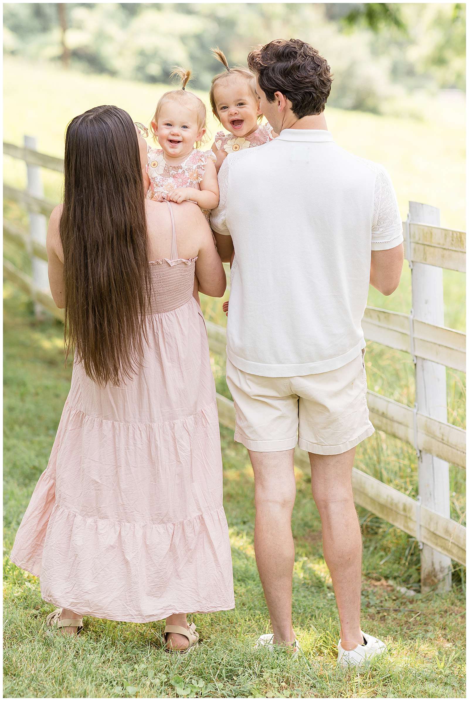 1 year old twins are held but their Mom and Dad who hold them over their shoulders and the twins look back and smile at Rebecca Rice Photography. They coordinate in outfits with blush, muted pinks, and white colors. -rebeccaricephoto.com
