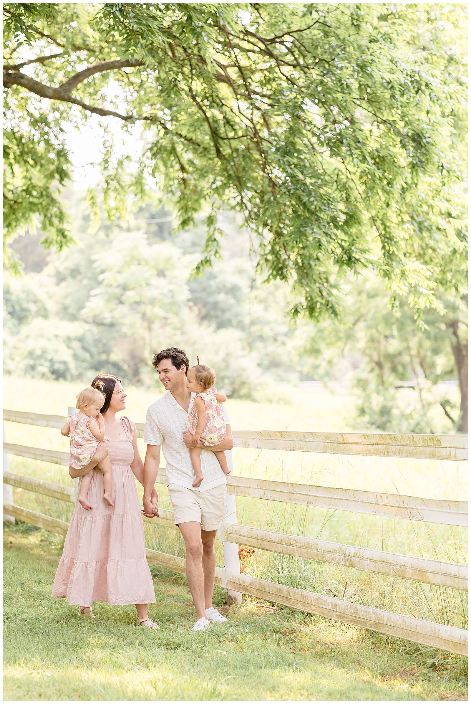 Mom and Dad hold hands and look at each other as they each hold their 1 year old twin daughters and walk along a white fence link surrounded by green grass and trees in Nashville for their family portraits! Click to see more photography education from Rebecca Rice Photography on the blog today! -rebeccaricephoto.com