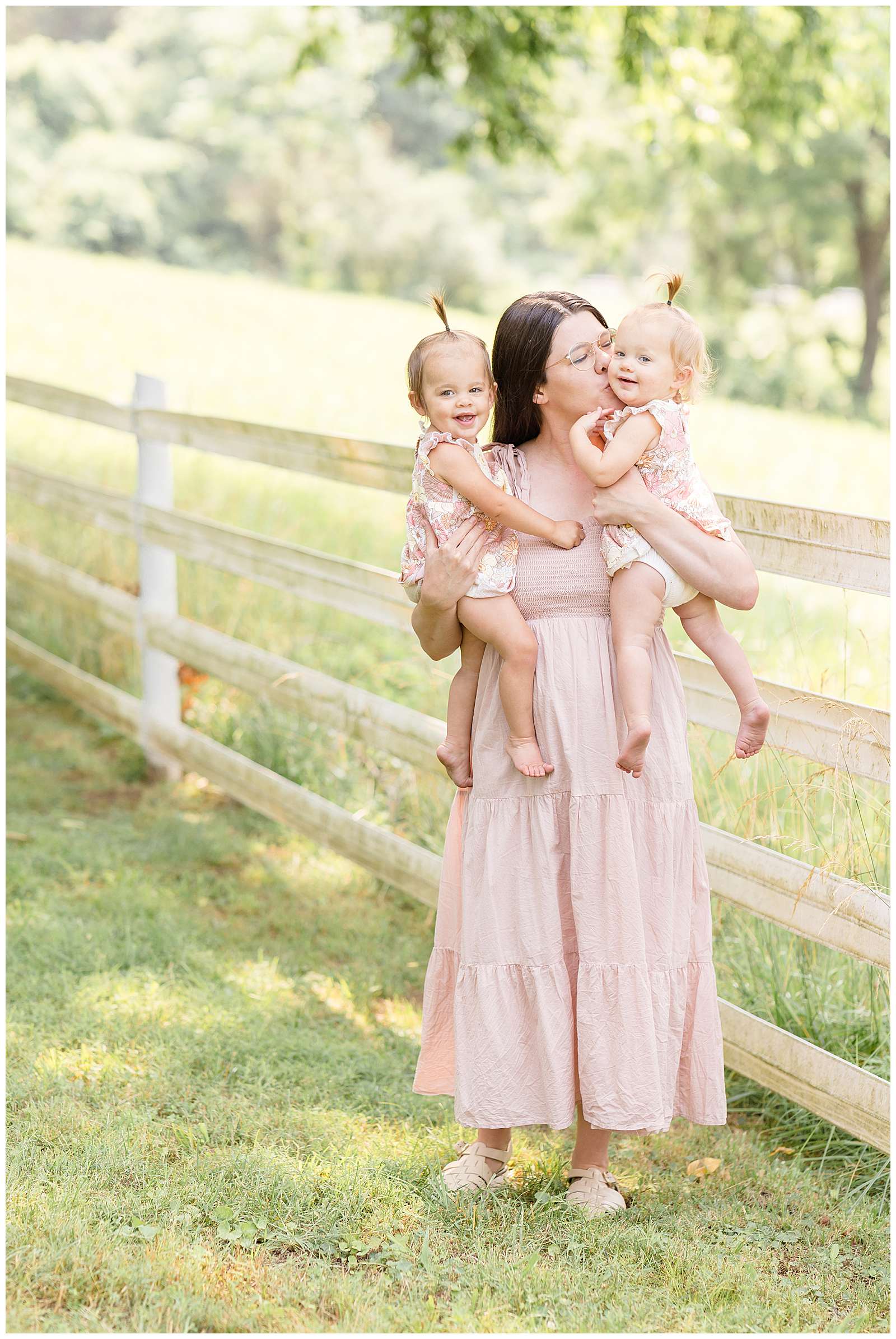 Sweet 1 year old twin girls who wear a neutral, muted pink tone floral onesies rompers with ruffle sleeves are head by their Mom who kisses one twin on the cheek during their Nashville family portrait session with Rebecca Rice Photography. Click to see more of these cuties on the blog today! -rebeccaricephoto.com
