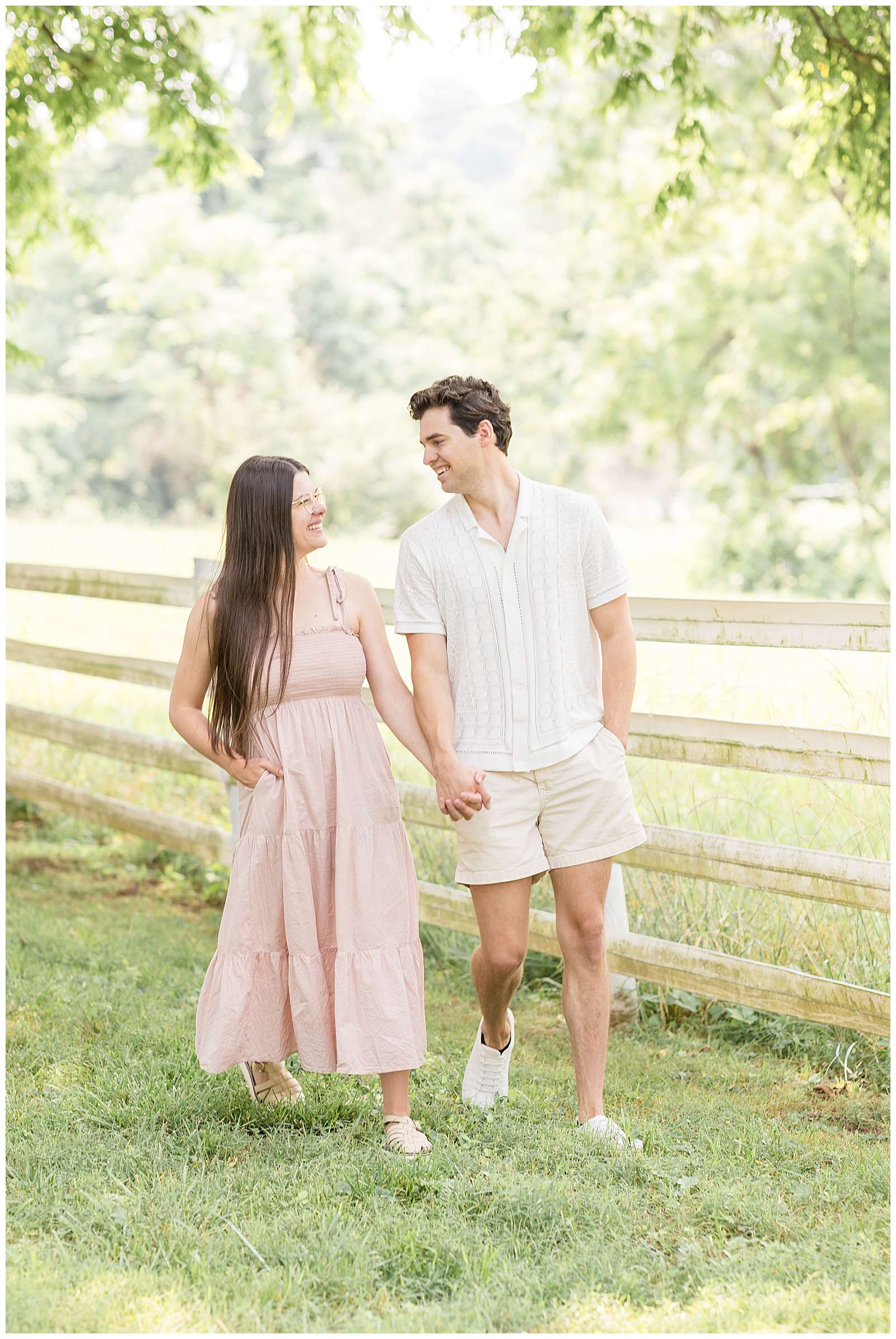 Young, new parents hold hands and walk together along a fence line smiling at each other. The girl wears a spaghetti strap blush, maxi dress, and her husband wears a white, button down shirt and light, khaki shorts. They walk together during Rebecca Rice's Behind the Lens Membership for her photography education! Check out this session and more about Behind the Lens on the blog today! -rebeccaricephoto.com
