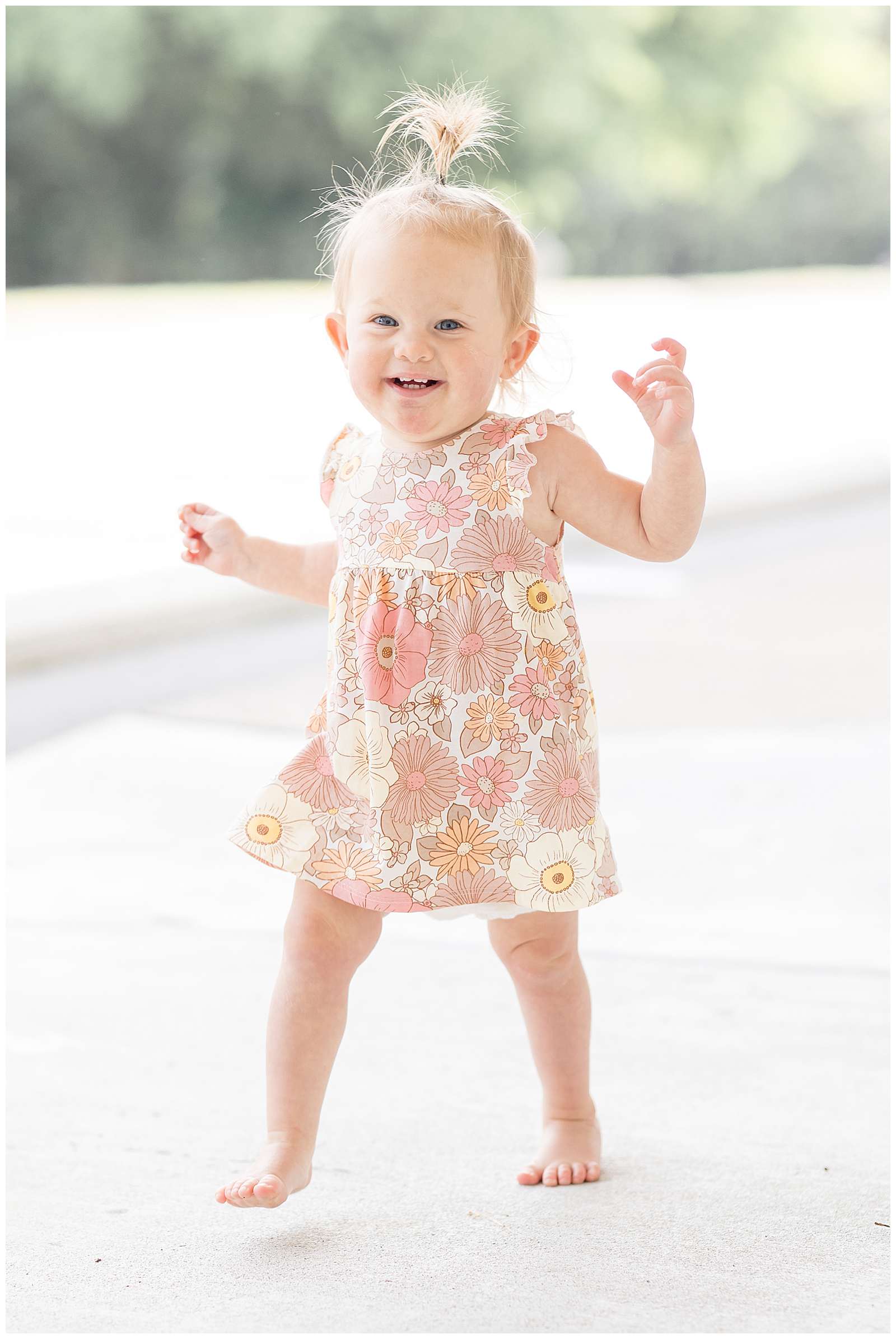 Sweet one year old girl with blonde hair and a whale sprout ponytail is a new walker and excited about taking her first steps. She smiles big at the camera and wears a muted, neutral floral dress with ruffle sleeves and no shoes. Click to see more of this cutie and her twin sister on the blog today! -rebeccaricephoto.com