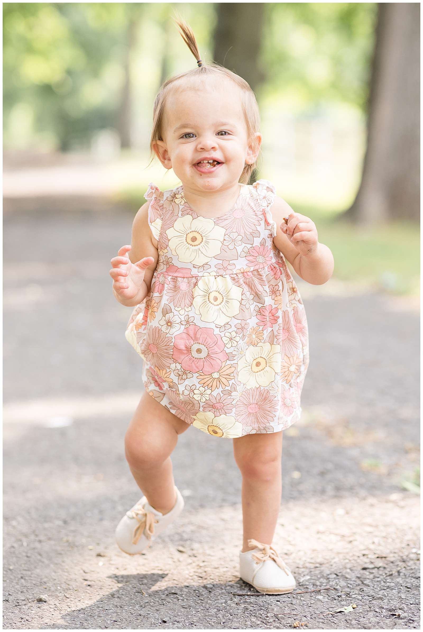 One year old girl walks along a pathway with a big grin and her tongue sticking through her teeth as she walks towards Rebecca Rice Photography. Click now to see more of this cutie and go behind the scenes with Rebecca Rice for her photography education membership. -rebeccaricephoto.com