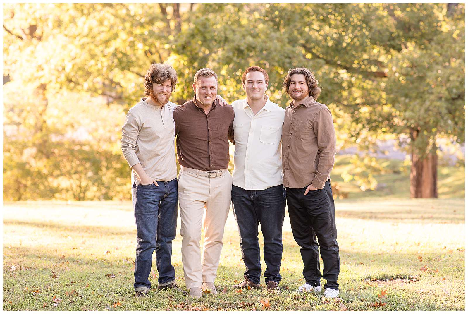 Dad stands in the middle wearing a brown button shirt and khakis as he stands with his arms around his grown 3 sons.  He has one on this left and the other 2 on his right.  The boys wear jeans and coordinating tones of tan.