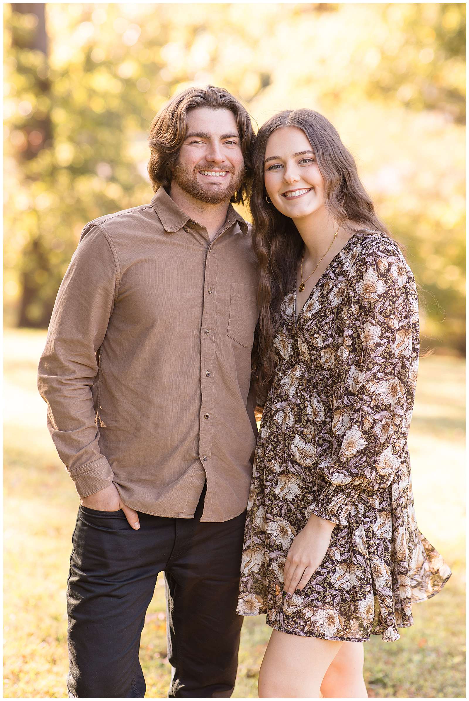 Young couple stands together during their fall family session in Tennessee.  They coordinate wearing brown tones with the boyfriend wearing a light brown button down shirt and dark pants and his girlfriend wears a brown dress with white and tan flowers on it.