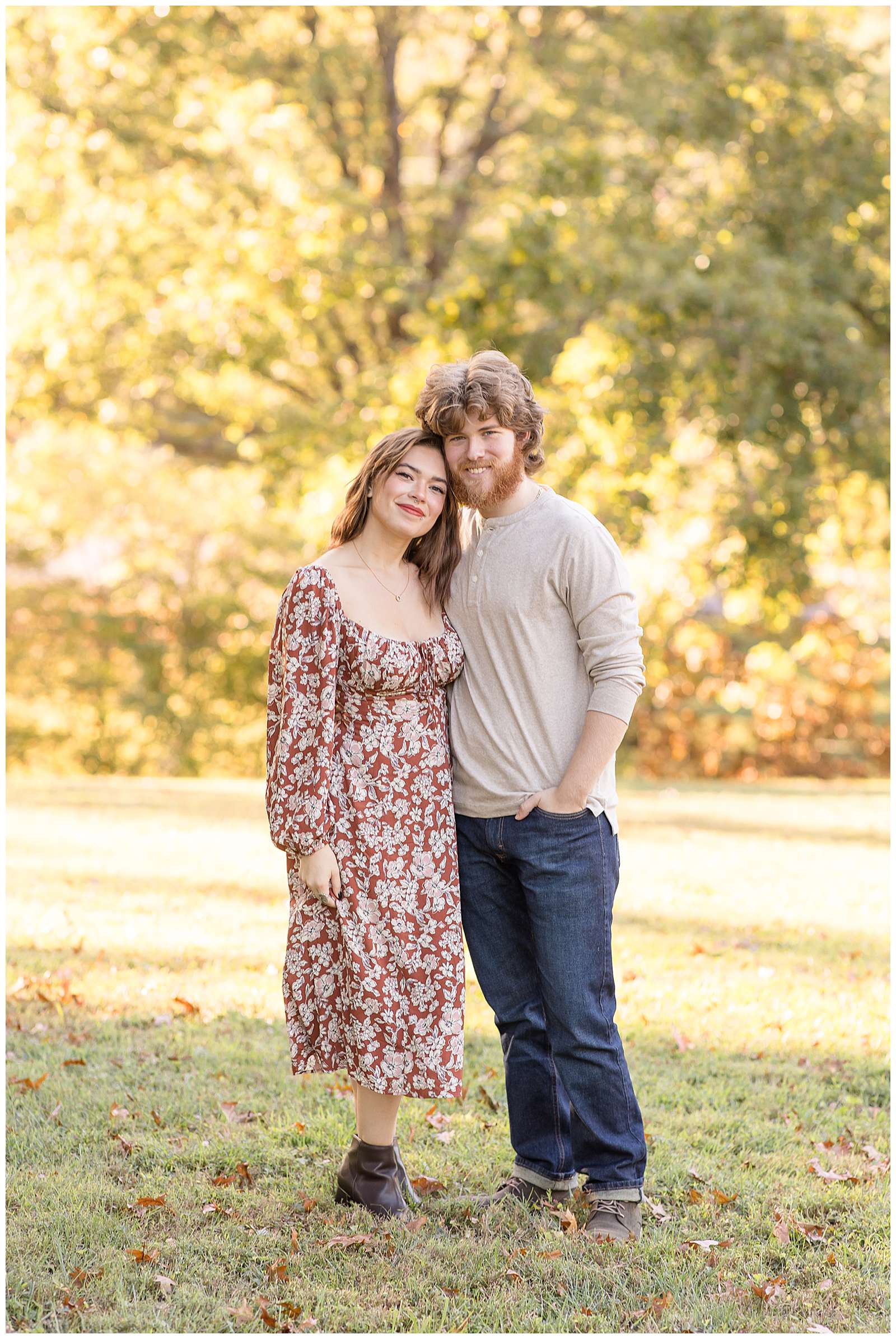 Couple stands together during their family fall portraits.  The 20 something girl wears an ankle length, long sleeve dress that is burnt orange with white flowers all over and her boyfriend wears jeans, and a tan, henley long sleeve shirt.