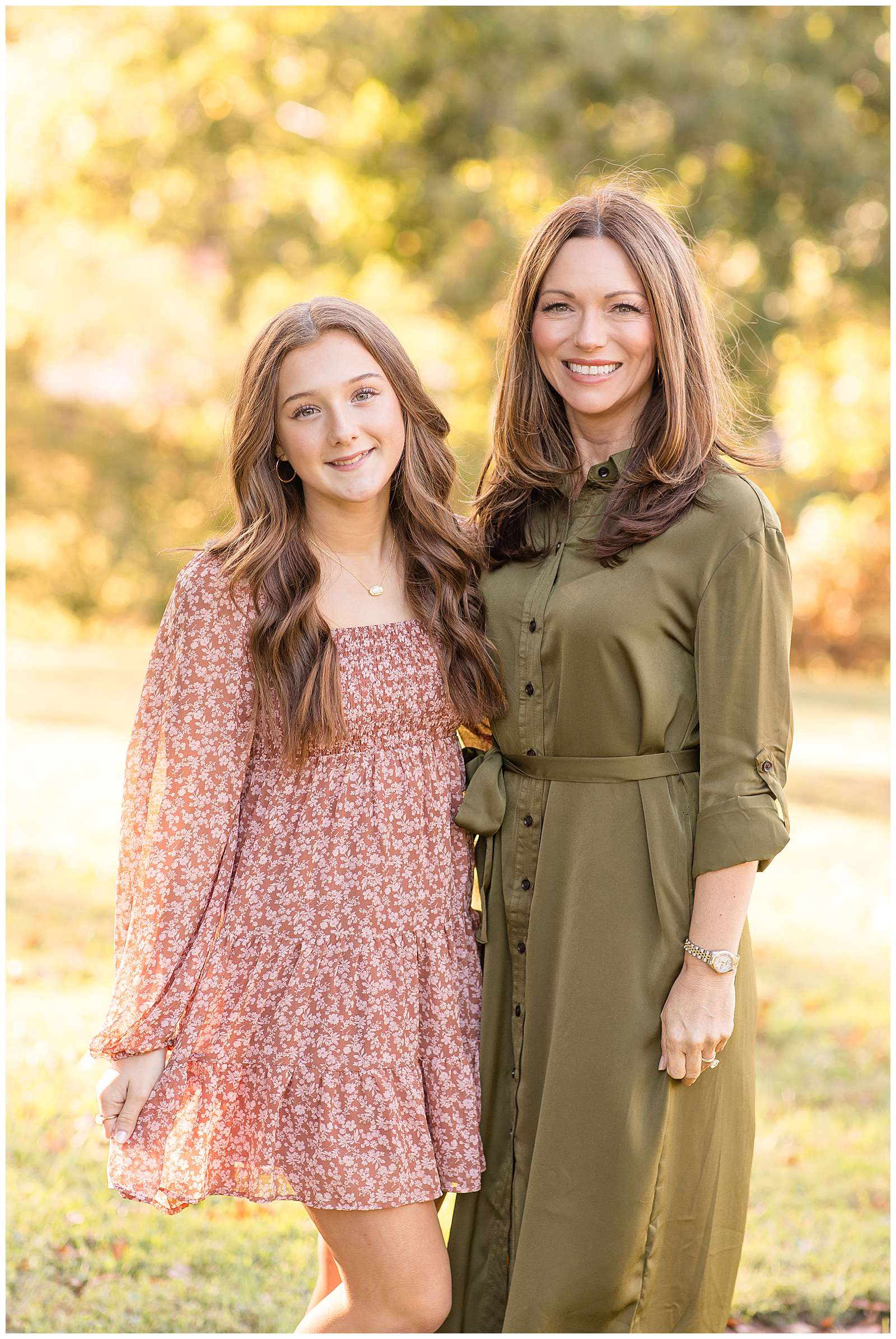 A mom who wears an olive green button down dress stands with her high school aged daughter who wears an above the knee, dusty pink, long sleeve dress with small white flower patterned dress.
